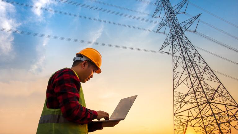 Power systems engineer working on a laptop beside electrical transmission infrastructure