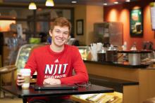 College student sitting in a coffee shop with a coffee cup and his laptop smiling wearing a NJIT shirt